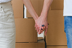 The hands of a woman and a man clasped firmly together as they stand before a stack of moving boxes.