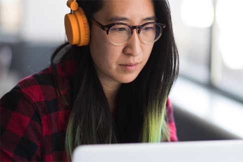 A woman with long, straight black hair, and headphones appears concentrated on the screen.
