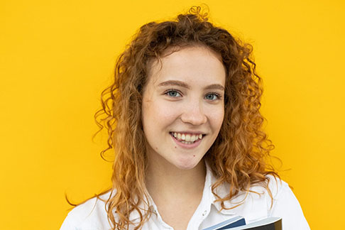 Against an orange-yellow background, a young woman with long, curly brown hair wears a cheerful smile.