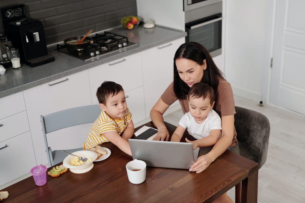 A young mother with two sons sits at a kitchen table, looking at a laptop computer.